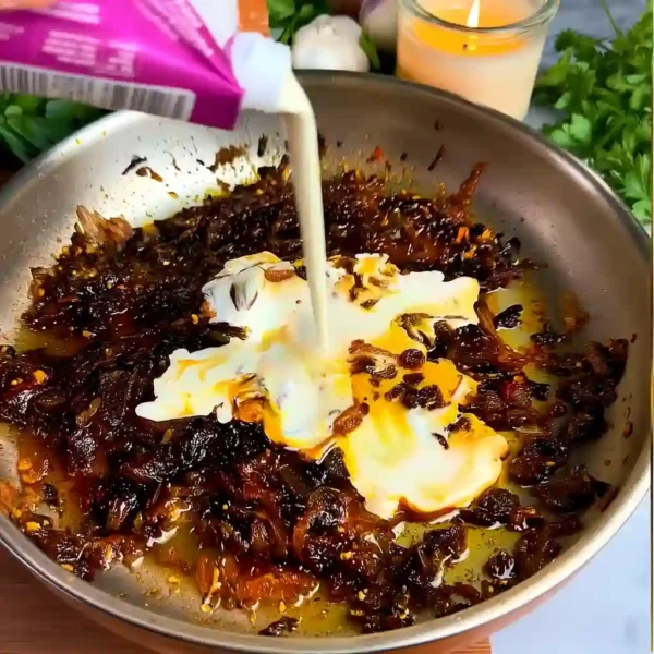 The Best Caramelized Onion and Garlic Spaghetti Cream being poured over caramelized onions and chili crisp in skillet.