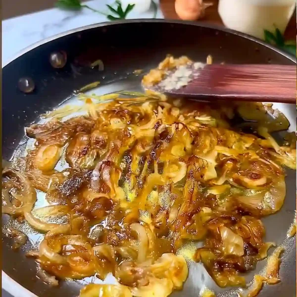 The Best Caramelized Onion and Garlic Spaghetti Golden caramelized onions cooking in a skillet with wooden spoon, soft candlelight background