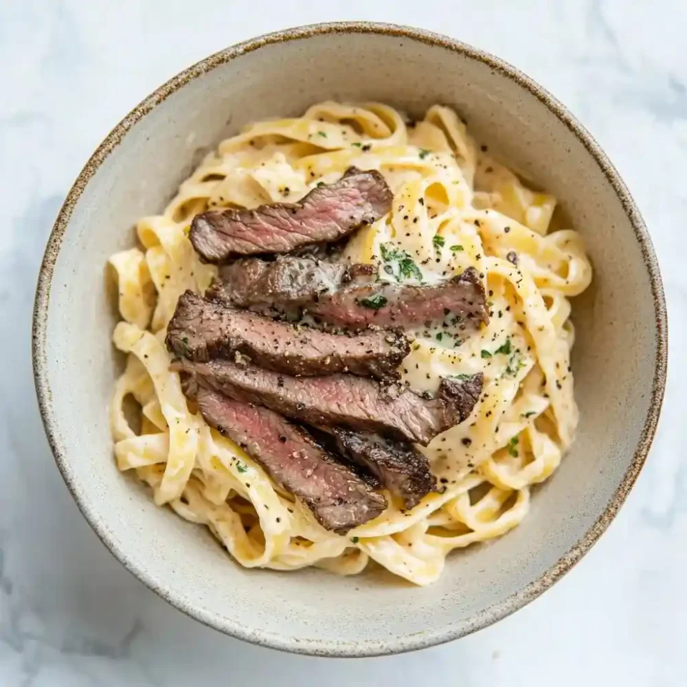 sliced beef over creamy alfredo pasta in a ceramic bowl