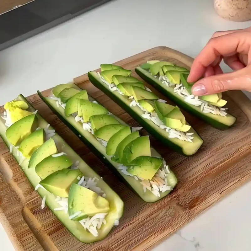 Hand assembling cucumber boats with rice and avocado slices on a wooden board.