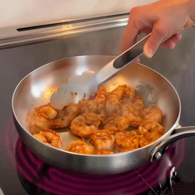 Hand using tongs to sauté shrimp in a steaming skillet on an electric stovetop